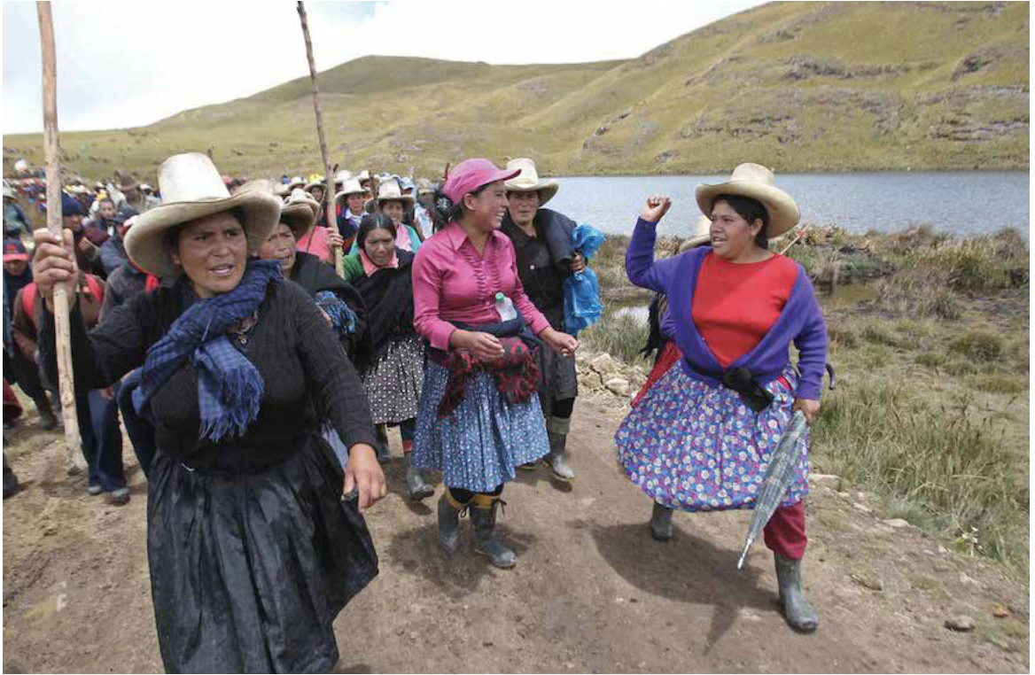 Group of indigenous women with sticks march in a hilly landscape.